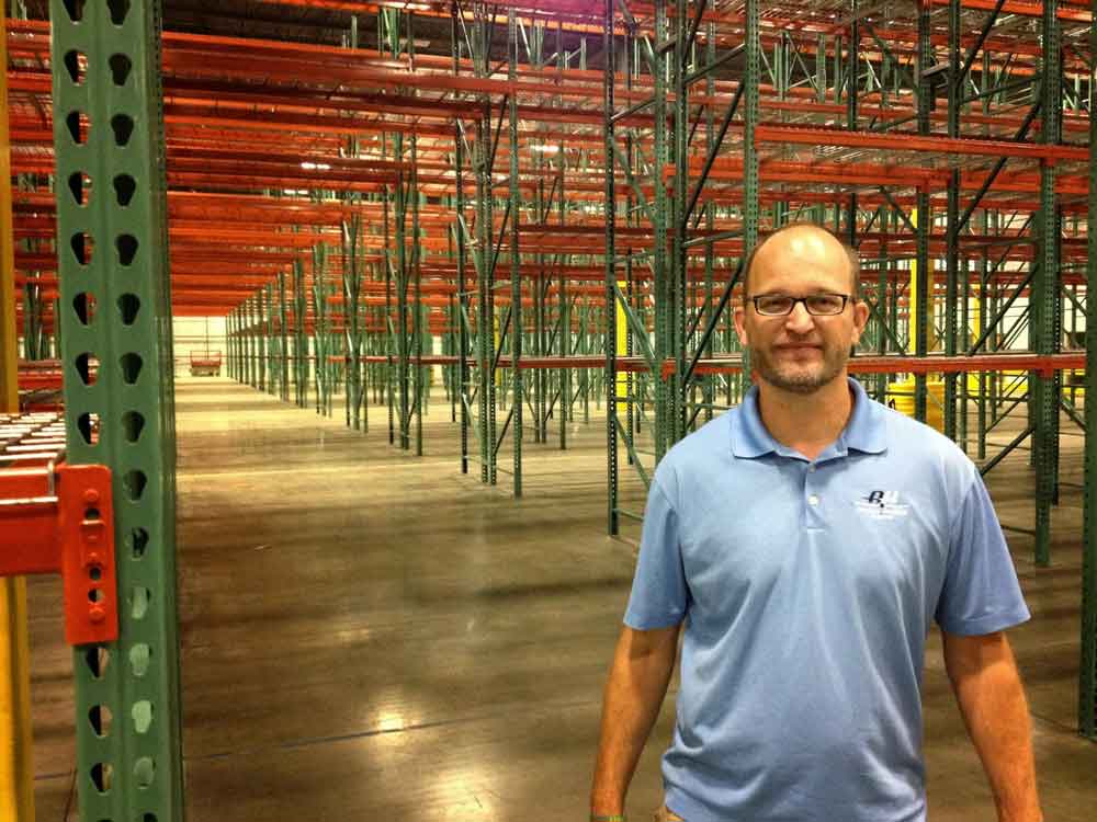 AKMHS Employee Brian Koski smiles while standing in front of empty bays of pallet racking in a warehouse