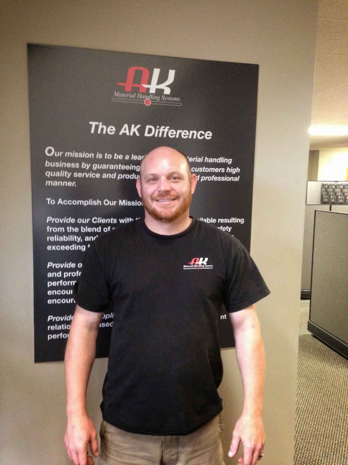 AKMHS Employee Tiege Quinn, smiles at the camera standing in front of a poster that outlines the company's values