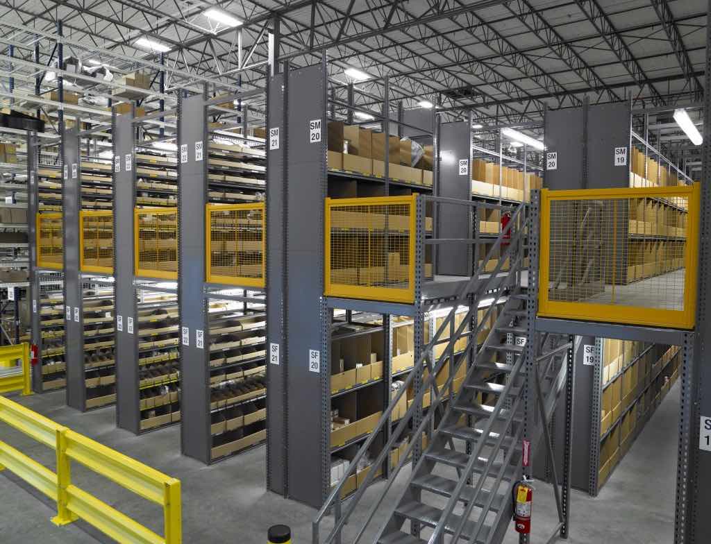 a stair case leads up to a mezzanine that has aisles of shelving storage above and below it