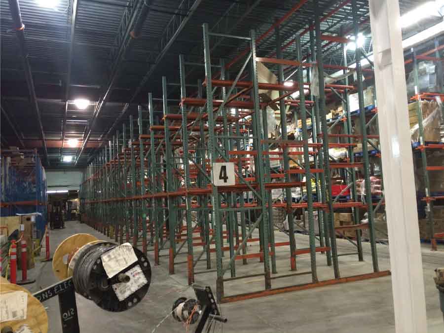 New racking stands in the Miner’s grocery store warehouse, behind it a row of pallet rack filled with boxes and other materials stands. A forklift a sits between blue and and green pallet racking on the other side